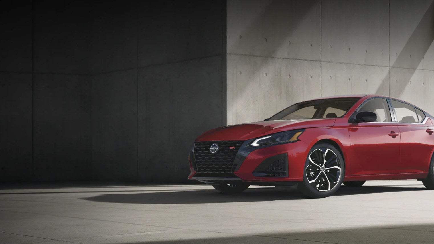 Red Nissan Altima sedan parked beside a concrete building with a city skyline behind it